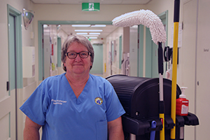 A woman stands in a corridor beside a trolley that holds cleaning equipment.