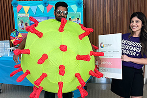 A man stands inside an inflatable suit resembling a germ. A woman stands beside him wearing a shirt that reads 'Antibiotic resistance fighter'