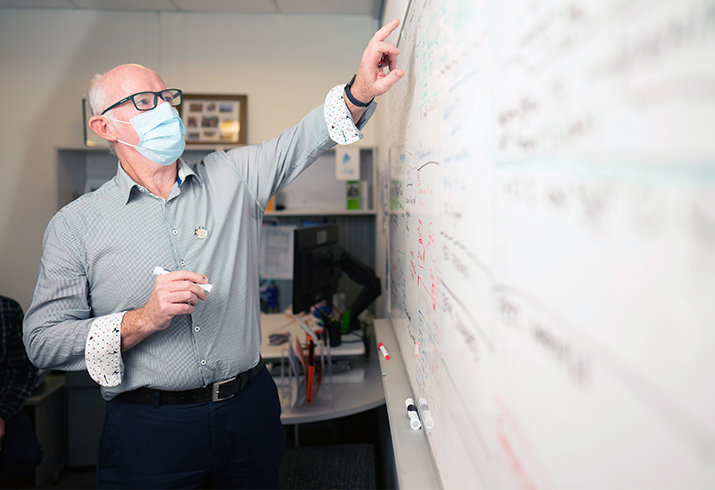 An older man wearing a surgical mask writes on a whiteboard.