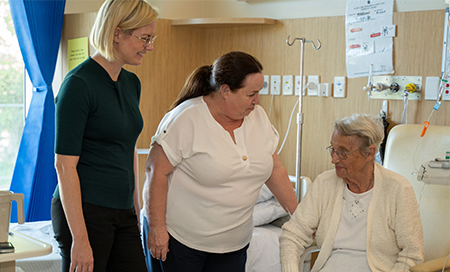 Two women stand talking to an older woman seated in a chair.