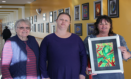 Three women stand in front of a series of artworks hanging on a bright yellow corridor wall. The woman on the right holds a framed painting of a frog.