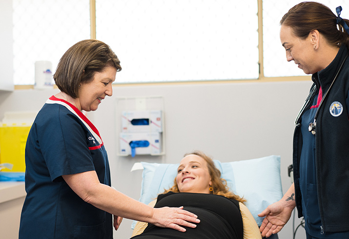 Two female midwives stand either side of a pregnant woman laying on a bed. One midwife has placed her hand on the women's stomach.