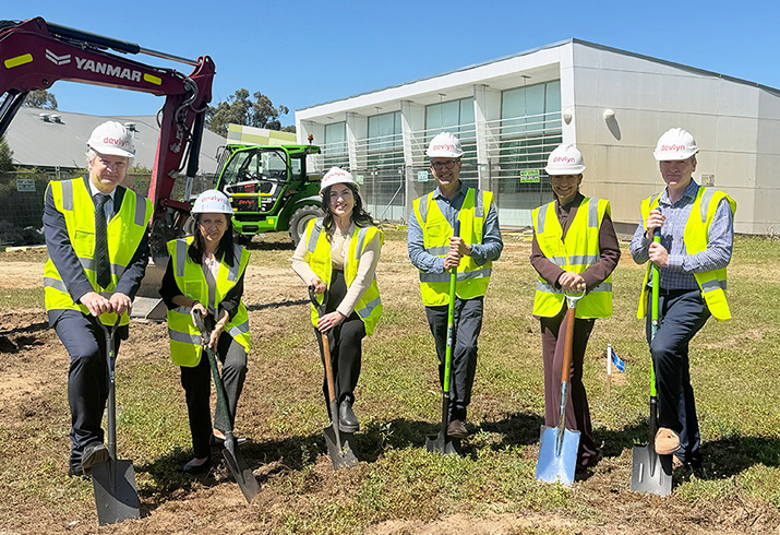 six people in safety gear holding shovels in front of a healthcare facility and construction equipment